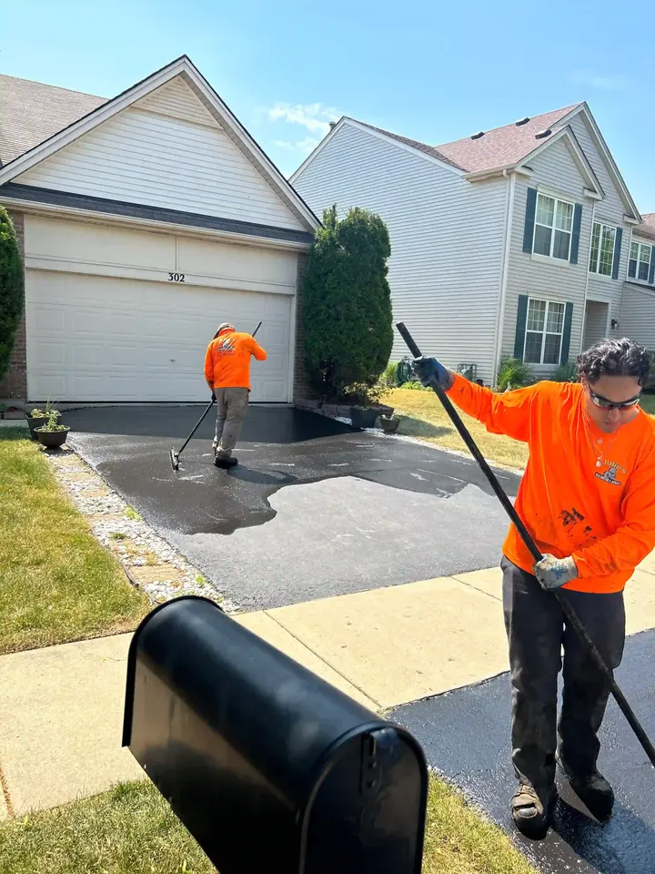 Crew members brushing sealcoat onto a driveway outside a light-colored house