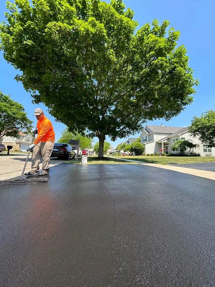 Worker applying sealcoat beneath a leafy tree beside a residential street
