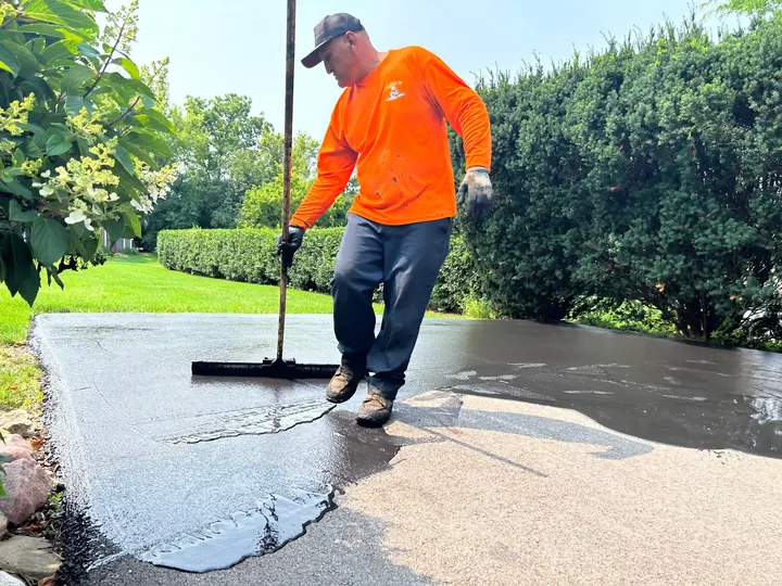 Worker spreading sealcoat with a wide brush near driveway landscaping