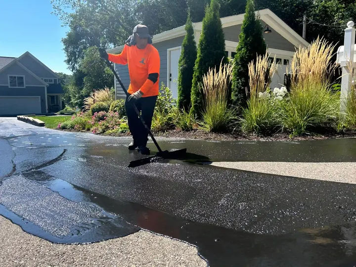 Worker brushing sealcoat onto a driveway beside ornamental grasses